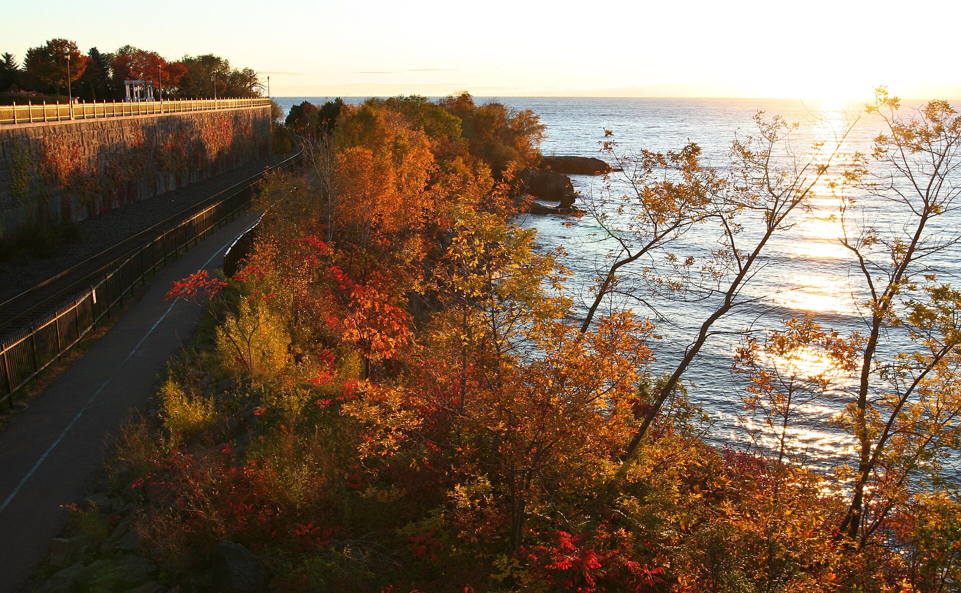 The paved Lakewalk running along the Lake Superior shoreline in Duluth below a stone retaining wall, with fall color on the bank