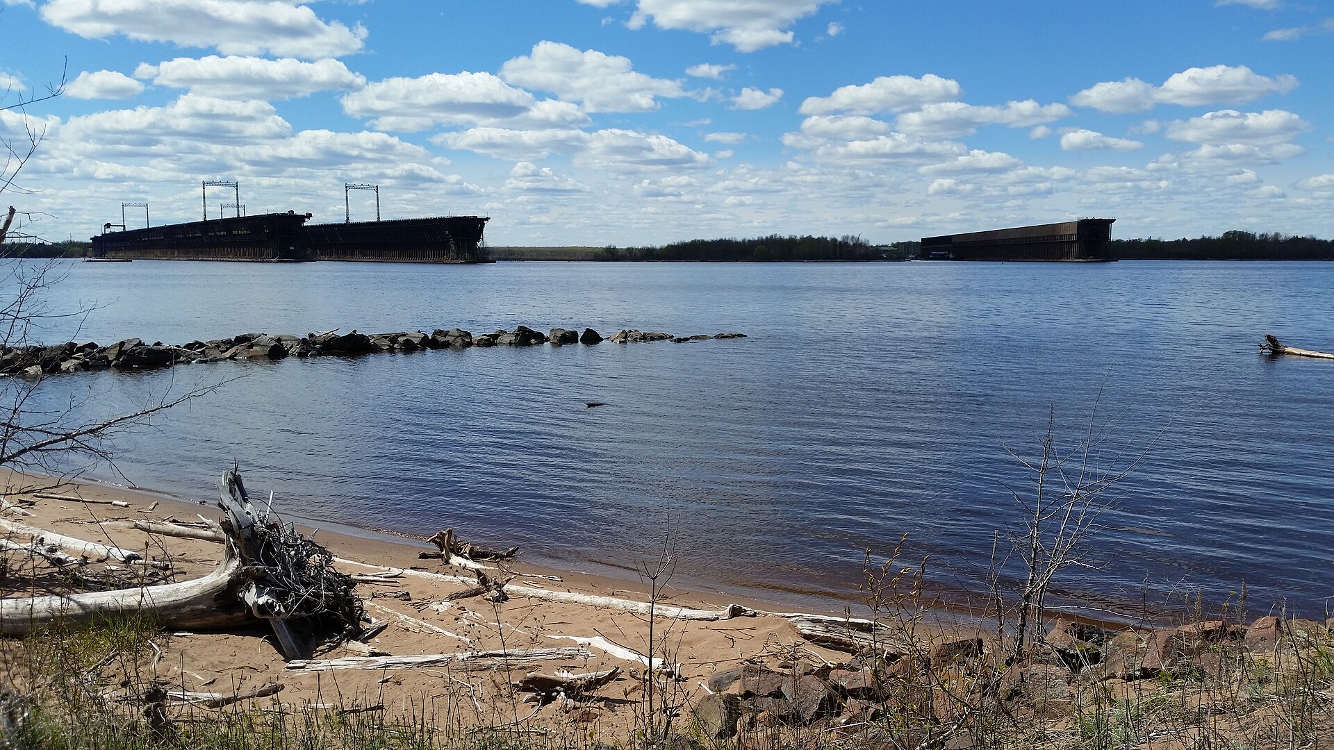 A sand and driftwood shoreline on Allouez Bay with the long low silhouettes of the Allouez ore docks across the water