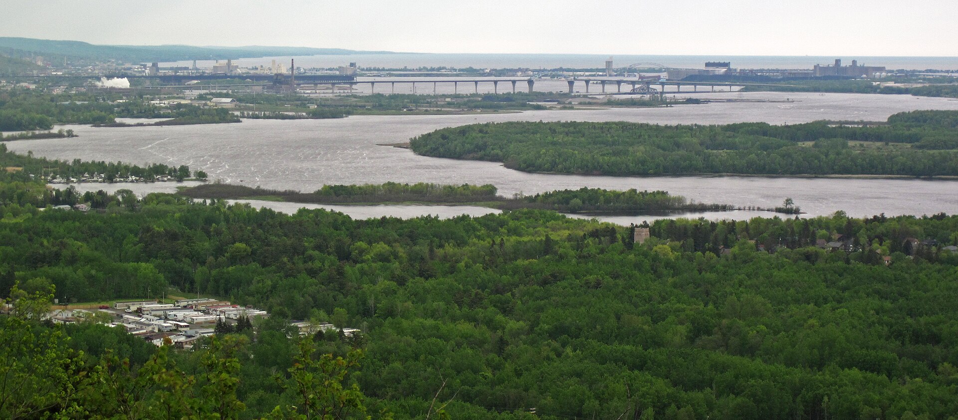 A wide view from the Duluth hilltop over wooded slopes to the St. Louis Bay, with the far shore and harbor industry beyond
