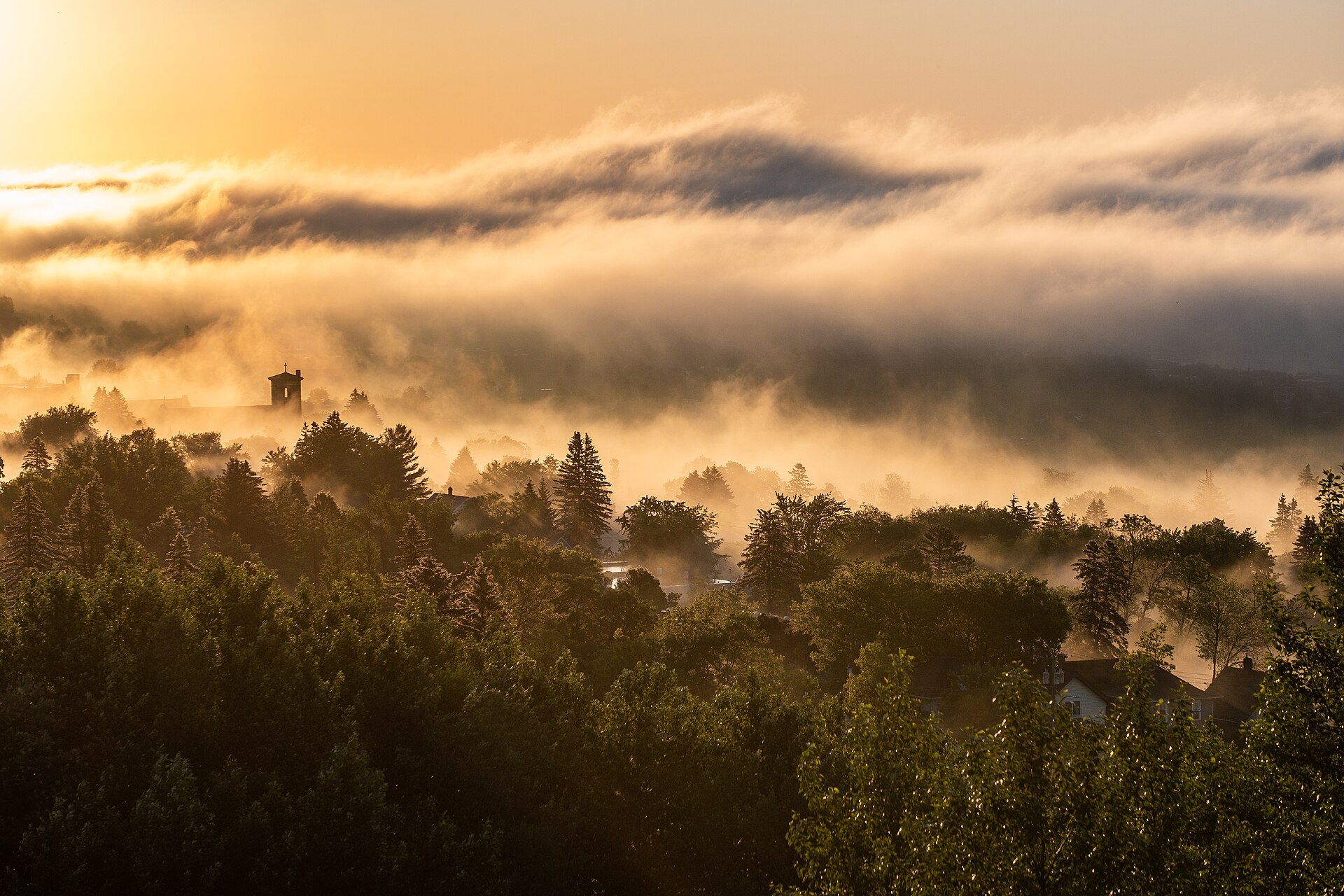 Morning fog burning off a wooded Duluth hillside at sunrise, with rooftops and a church tower showing through the trees