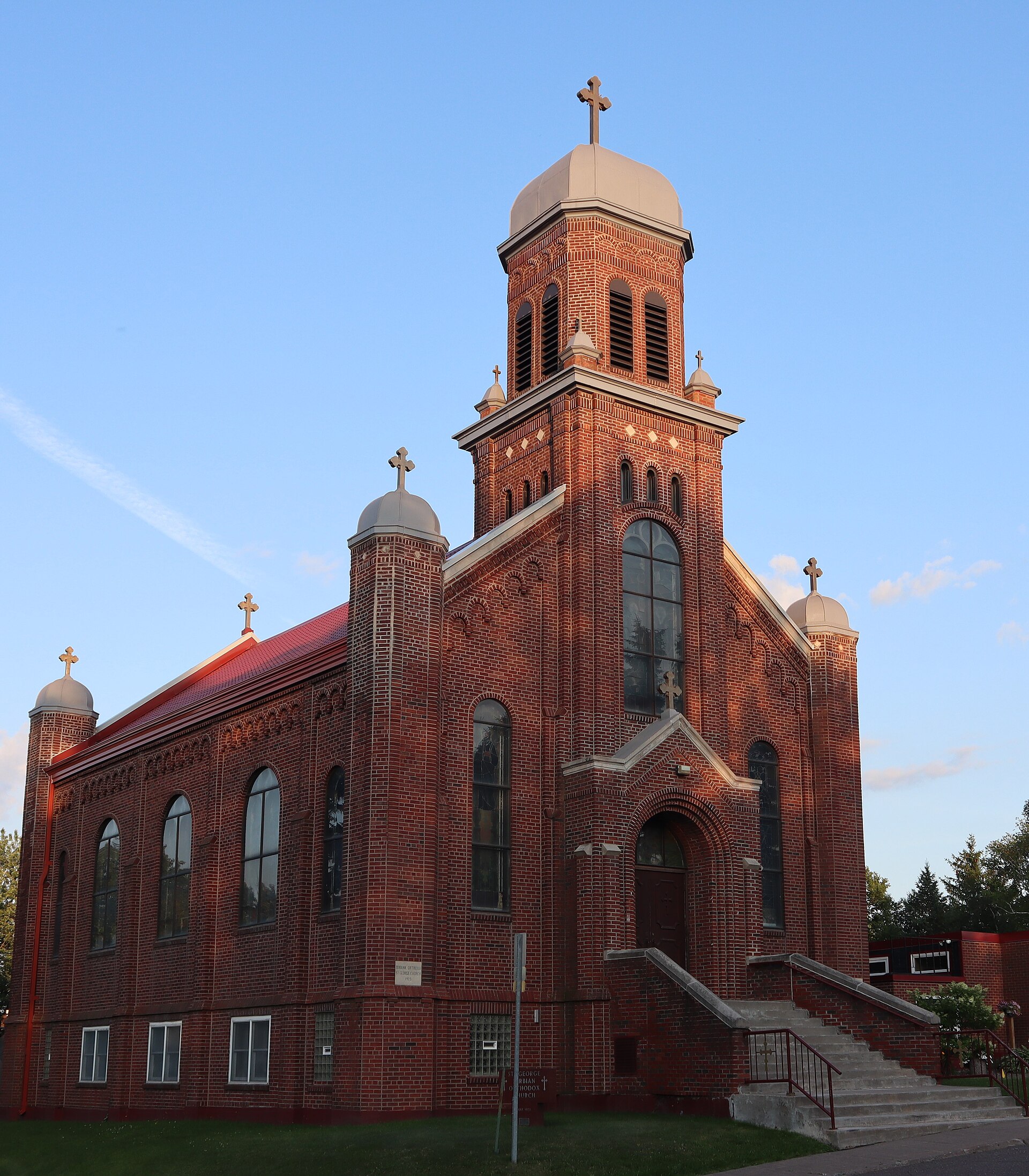 A red brick Serbian Orthodox church with a domed bell tower and silver cupolas, lit by low evening sun in Gary-New Duluth