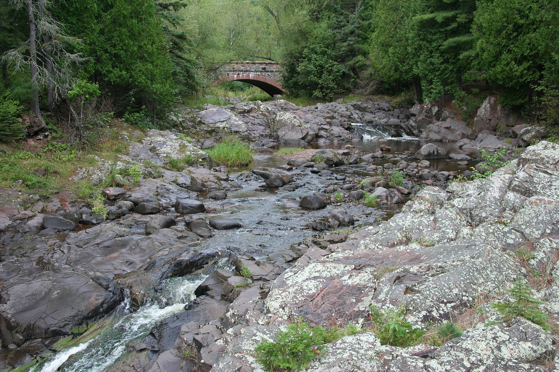 Amity Creek running over bare rock ledges toward a stone arch footbridge in Lester Park, Duluth