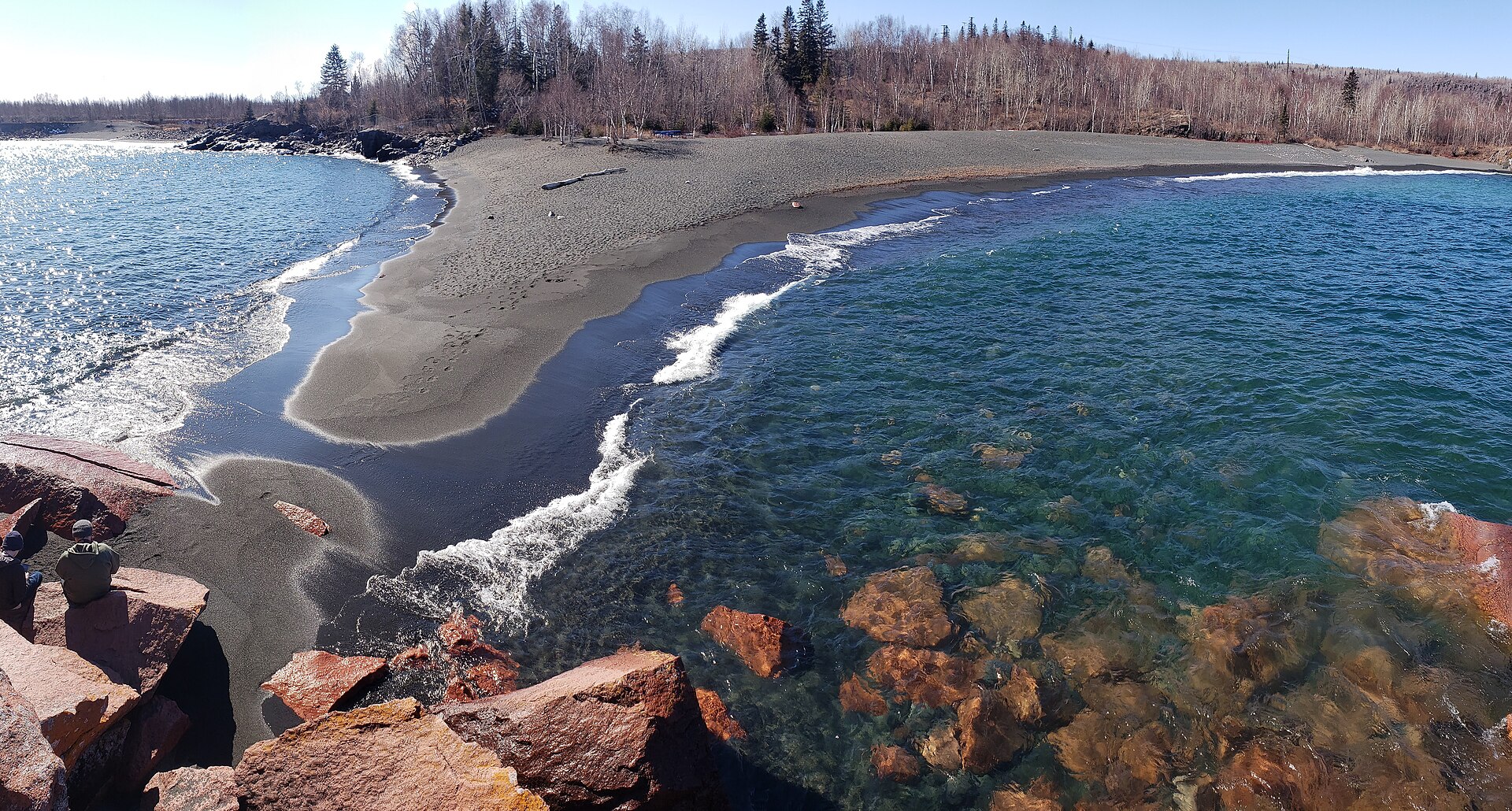A dark grey taconite sand beach curving between red rock and clear shallow water on Lake Superior at Silver Bay