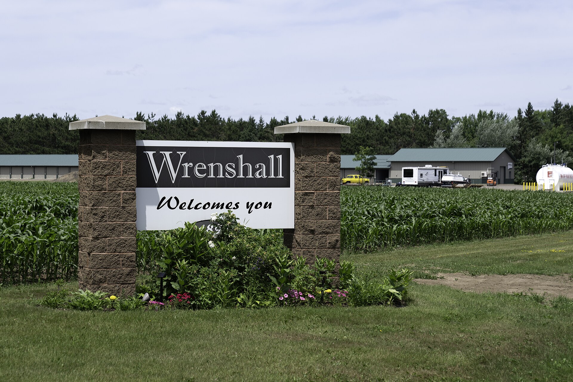 A stone and painted sign reading Wrenshall Welcomes You, set in flowers at the edge of a green corn field